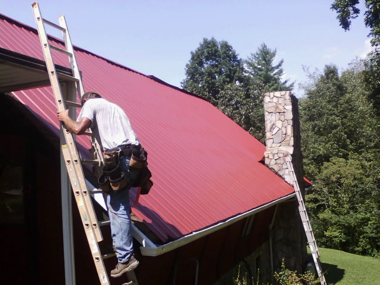 Roofer on a ladder beside a red metal roof and stone chimney — Soffit & Fascia Repair by Roofing Brothers in New Ringgold